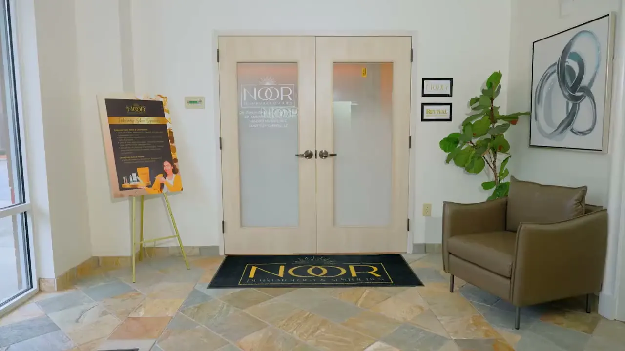 The entrance to NOOR Dermatology & Aesthetics: frosted double doors etched with the NOOR logo, a gold-lettered NOOR doormat, a specials poster on an easel, and a leather armchair beside a potted plant.