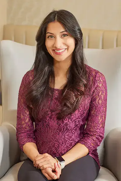 Dr. Syeda Amna Husain, smiling, seated in an armchair in the NOOR Dermatology & Aesthetics office.