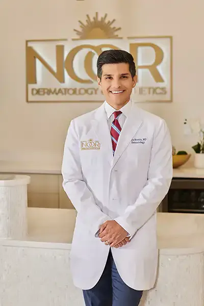 Dr. Zain Husain in a white coat, standing in the NOOR Dermatology & Aesthetics reception in front of the gold NOOR logo.