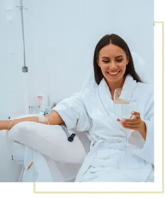 A smiling woman in a white robe reclining in a treatment chair with an IV line in her arm, holding a drink, an IV stand beside her.