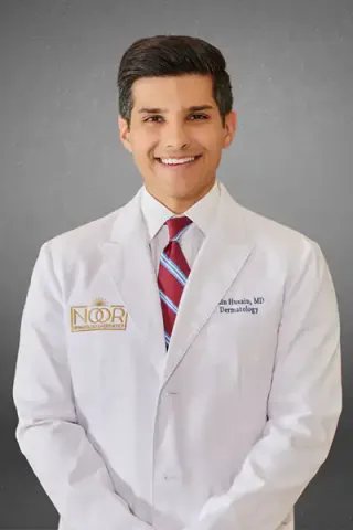 Dr. Zain Husain, founder of NOOR Dermatology & Aesthetics, in a white lab coat embroidered with the NOOR logo and his name, smiling against a mottled grey studio background.